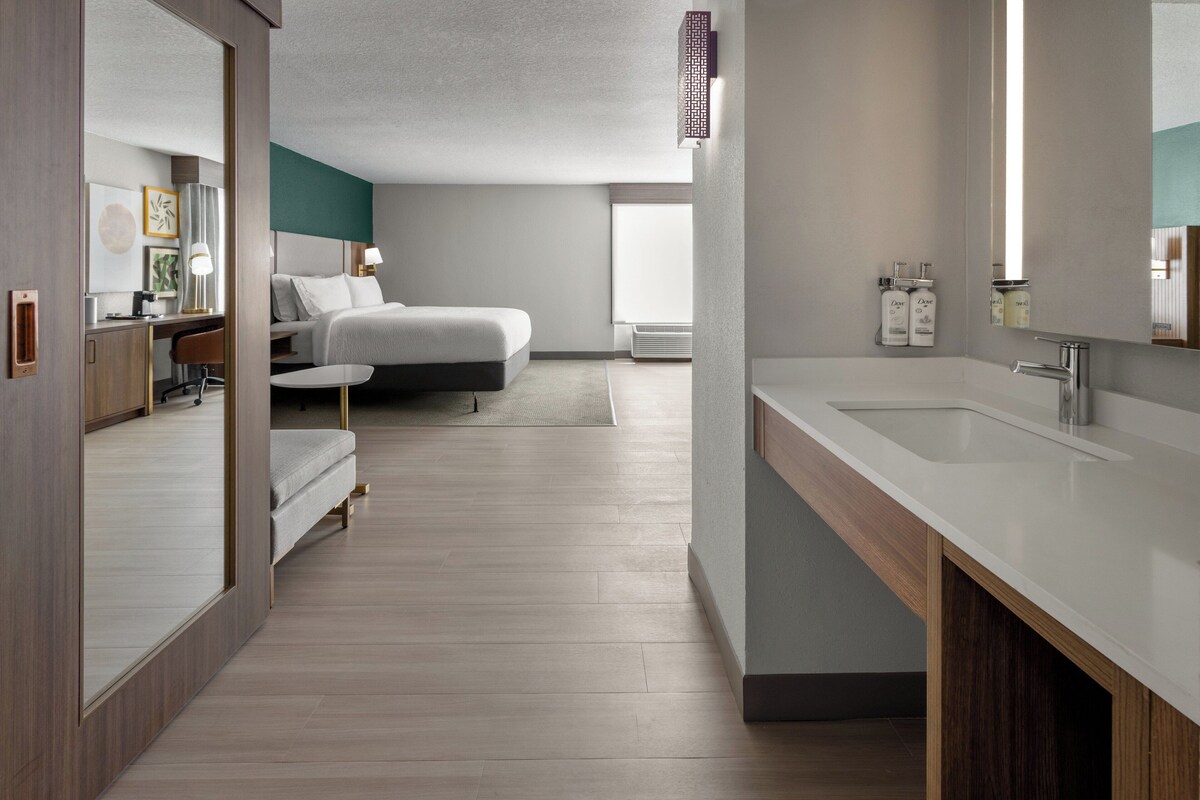 Una habitación de hotel moderna con una cama grande, una zona de estar con escritorio y silla, un baño amplio con lavamanos y espejo, y vistas al exterior.