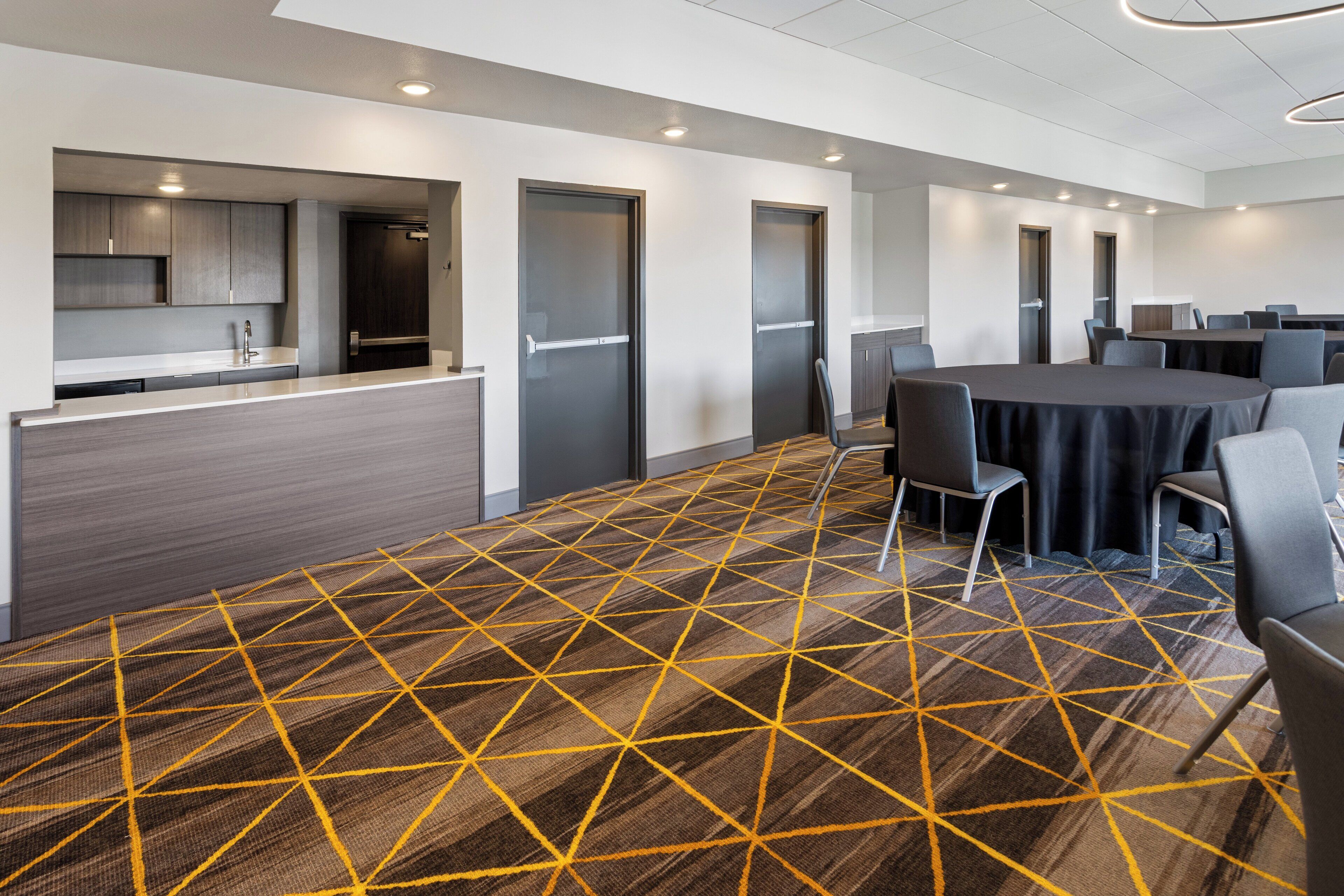 A modern conference room with a geometric patterned carpet, a long table with chairs, and a kitchenette area with cabinets and a sink.