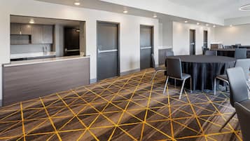 A modern conference room with a geometric patterned carpet, a long table with chairs, and a kitchenette area with cabinets and a sink.