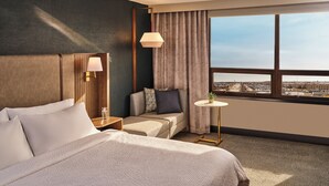 Habitación de hotel con una cama grande, una mesita de noche con lámpara, una pequeña mesa con planta, un sofá con cojines y vistas a la ciudad a través de un ventanal.