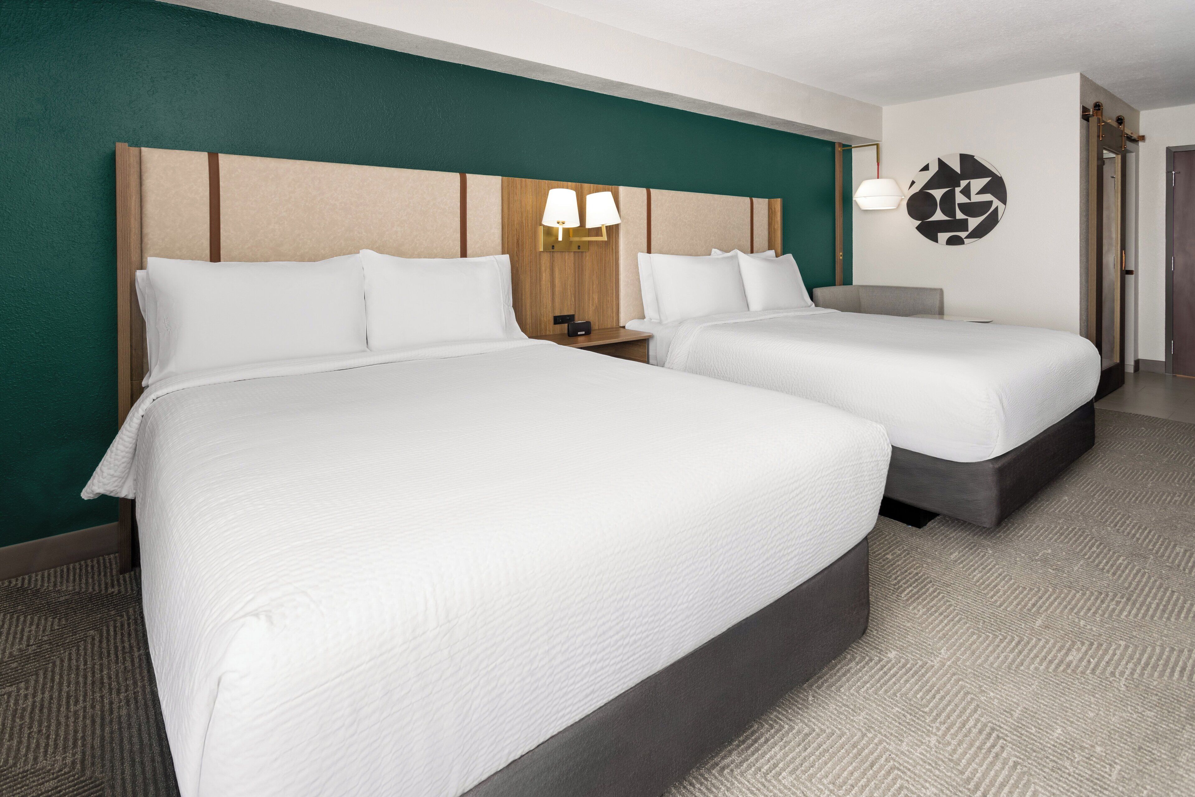 A hotel room with two beds, a headboard, bedside lamps, a wall art piece, and a carpeted floor.