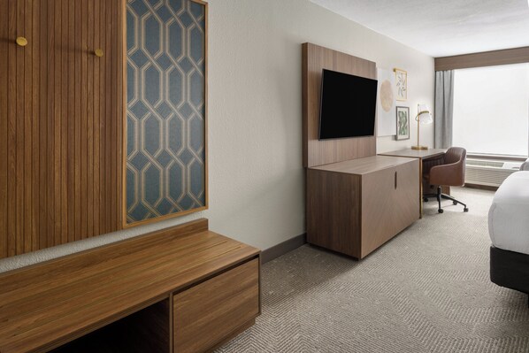 A hotel room with a wooden wardrobe, a desk with a chair, a flat-screen TV, and a large window providing a view of the city.