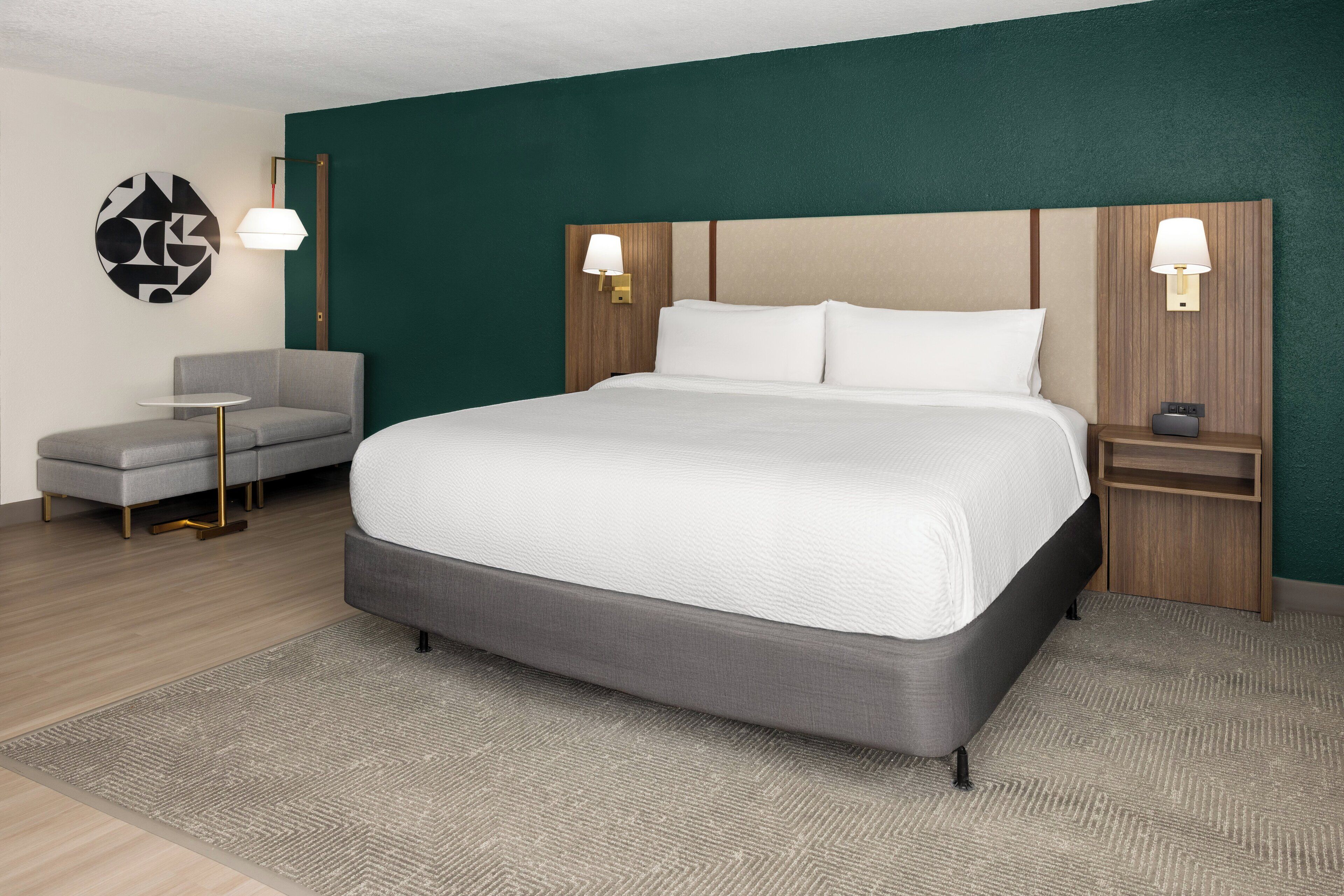 Une chambre d’hôtel moderne avec un grand lit, deux tables de chevet avec des lampes, un fauteuil gris et une petite table ronde.