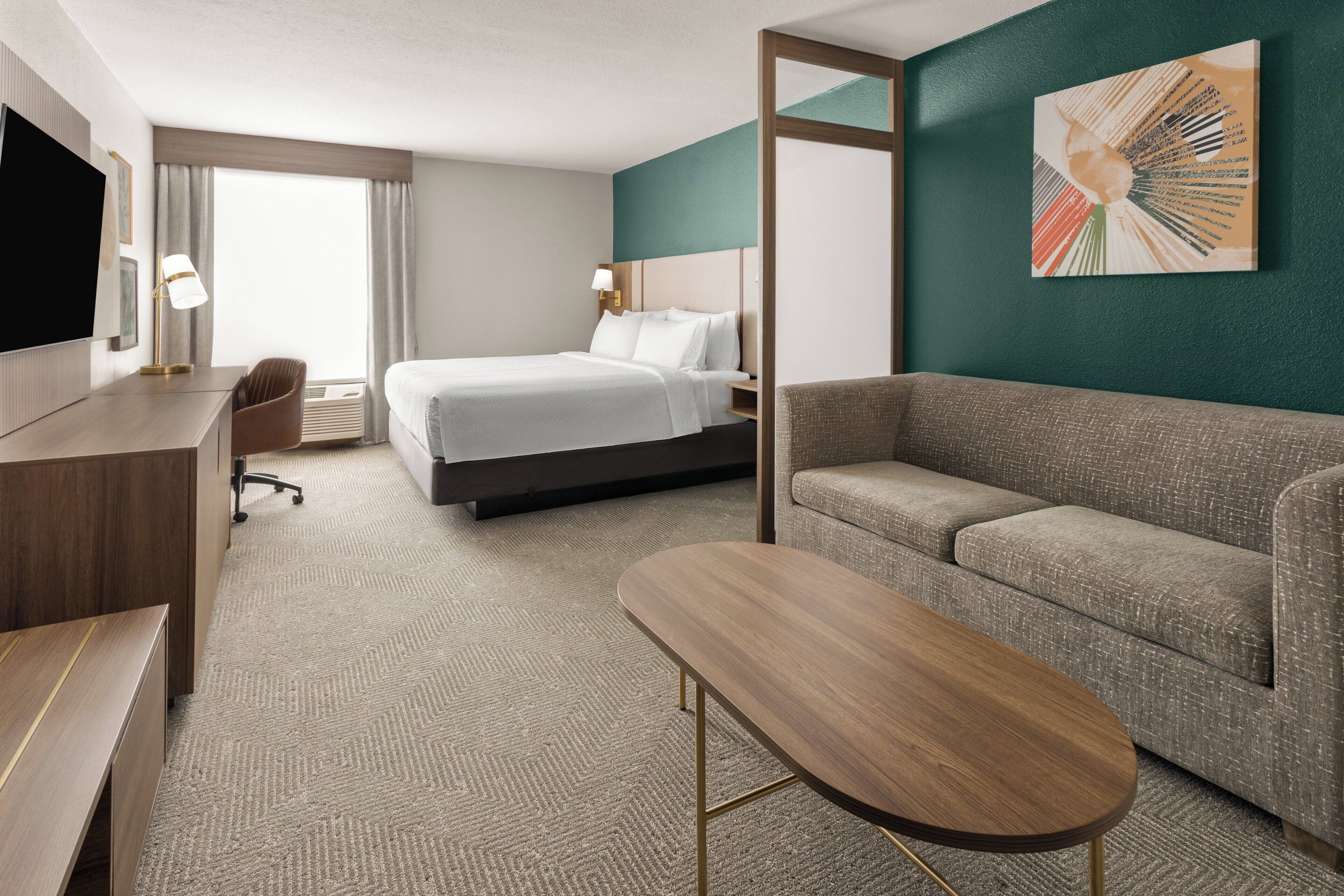 A hotel room with a bed, a sofa, a desk, a TV, and a painting on the wall.