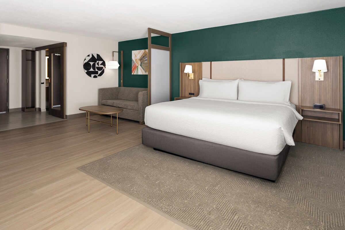 A modern hotel room with a large bed, a sofa, a small table, and a wall-mounted lamp.