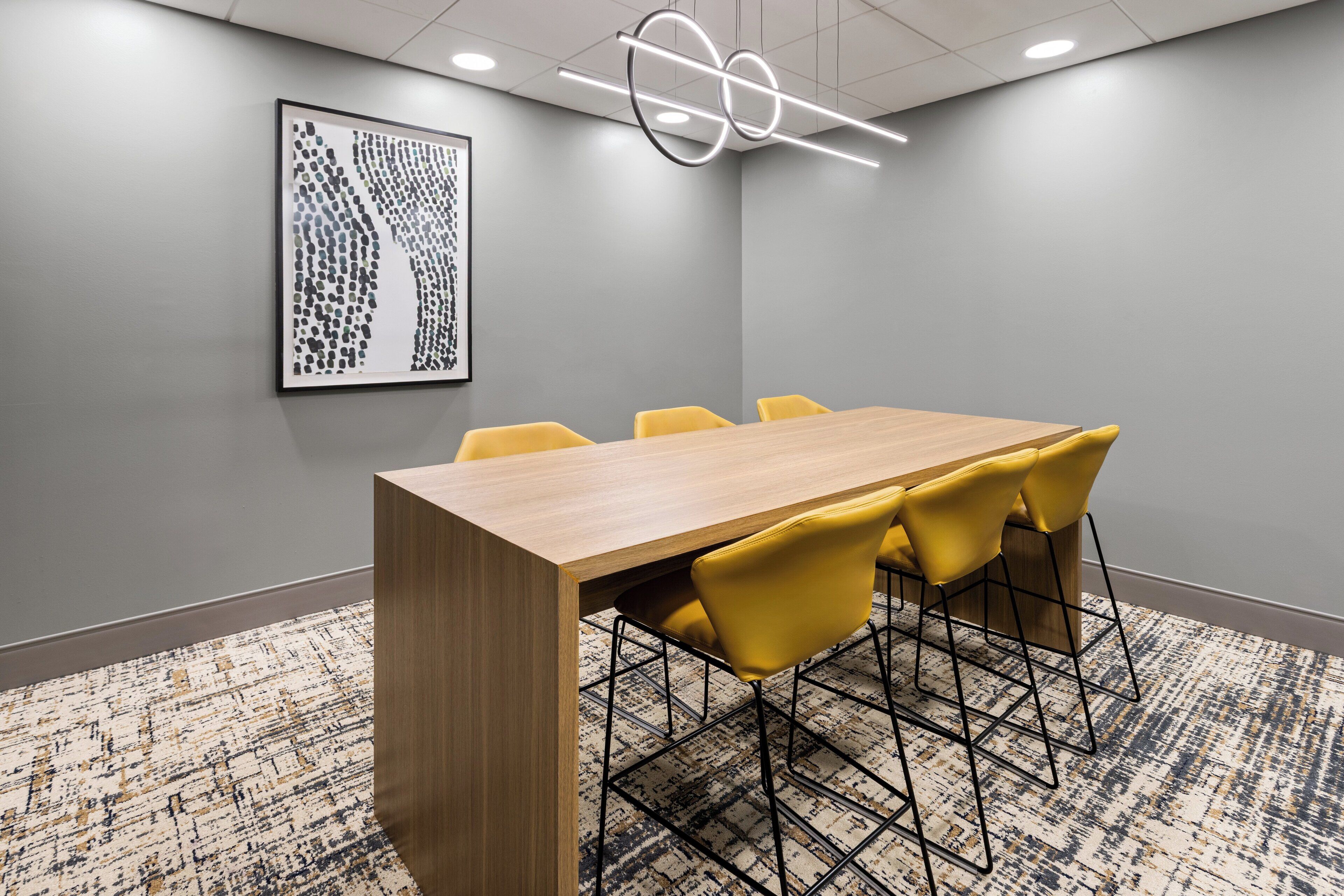 A conference room with a wooden table, yellow chairs, a patterned carpet, and a framed artwork on the wall.