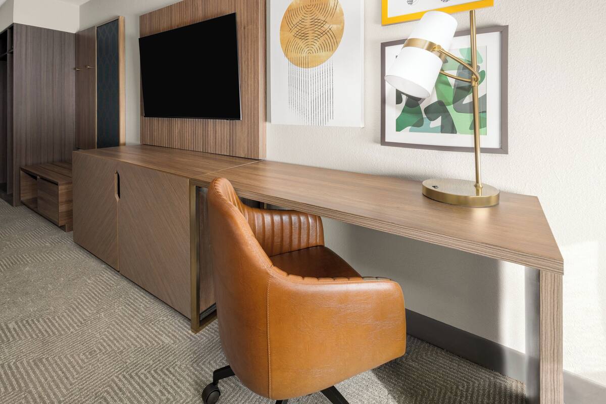 A modern hotel room with a wooden desk, a leather chair, a flat-screen TV, and a framed abstract painting.