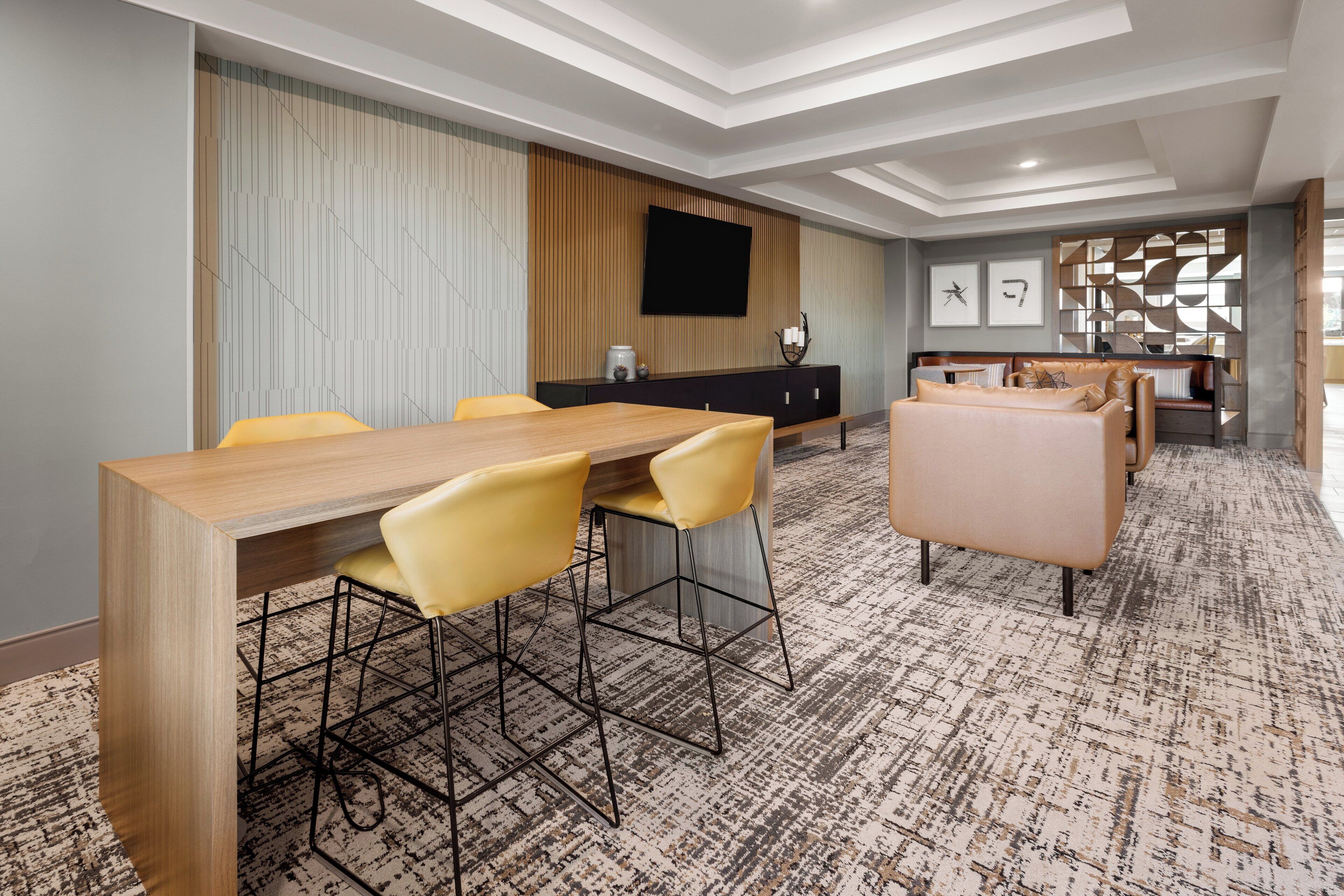A modern hotel room with a wooden dining table, yellow chairs, a sofa, and a TV mounted on the wall.