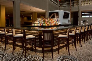 Zona de bar con una barra central que exhibe diversas botellas, rodeada de taburetes.