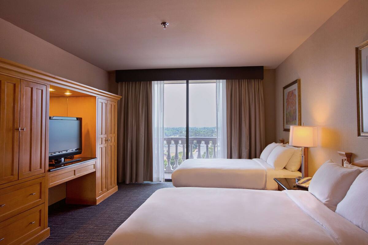 A hotel room with two beds, a TV, a desk, and a balcony with curtains.