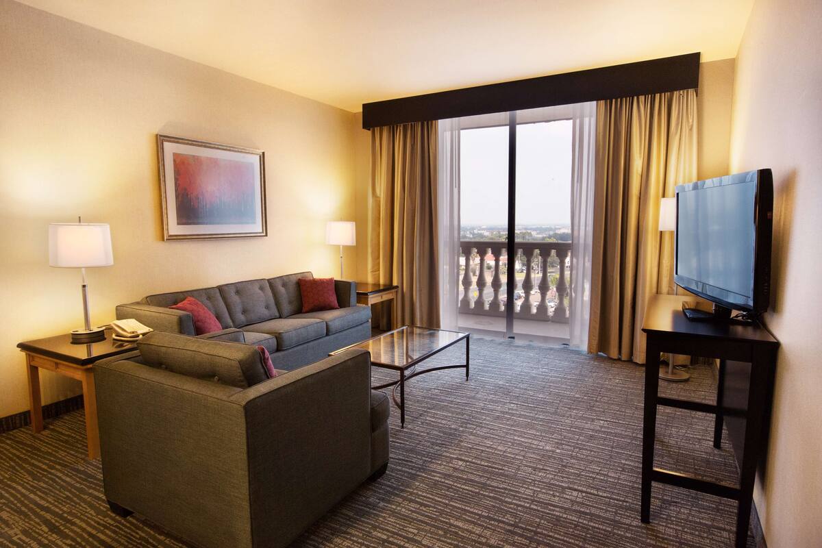 A hotel room with a sofa, a coffee table, a TV on a stand, and a balcony with curtains.