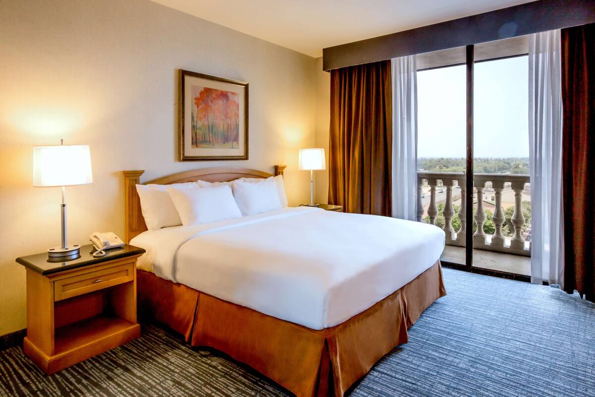 A hotel room with a large bed, two bedside tables with lamps, a painting on the wall, and a balcony with a view.