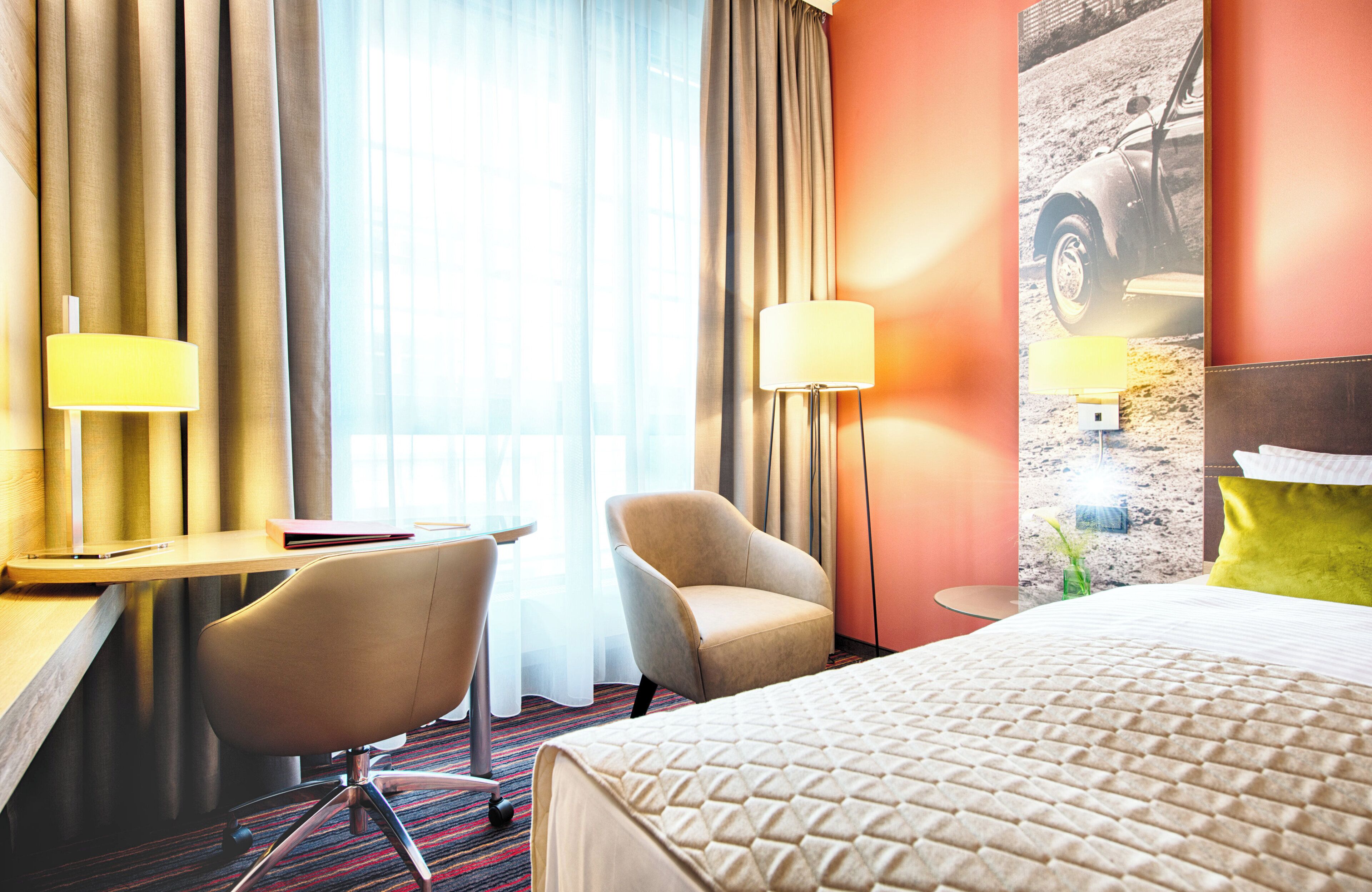 Hotellihuone, jossa on sänky, kaksi tuolia, työpöytä, lamppu ja suuri ikkuna, jossa on verhot.