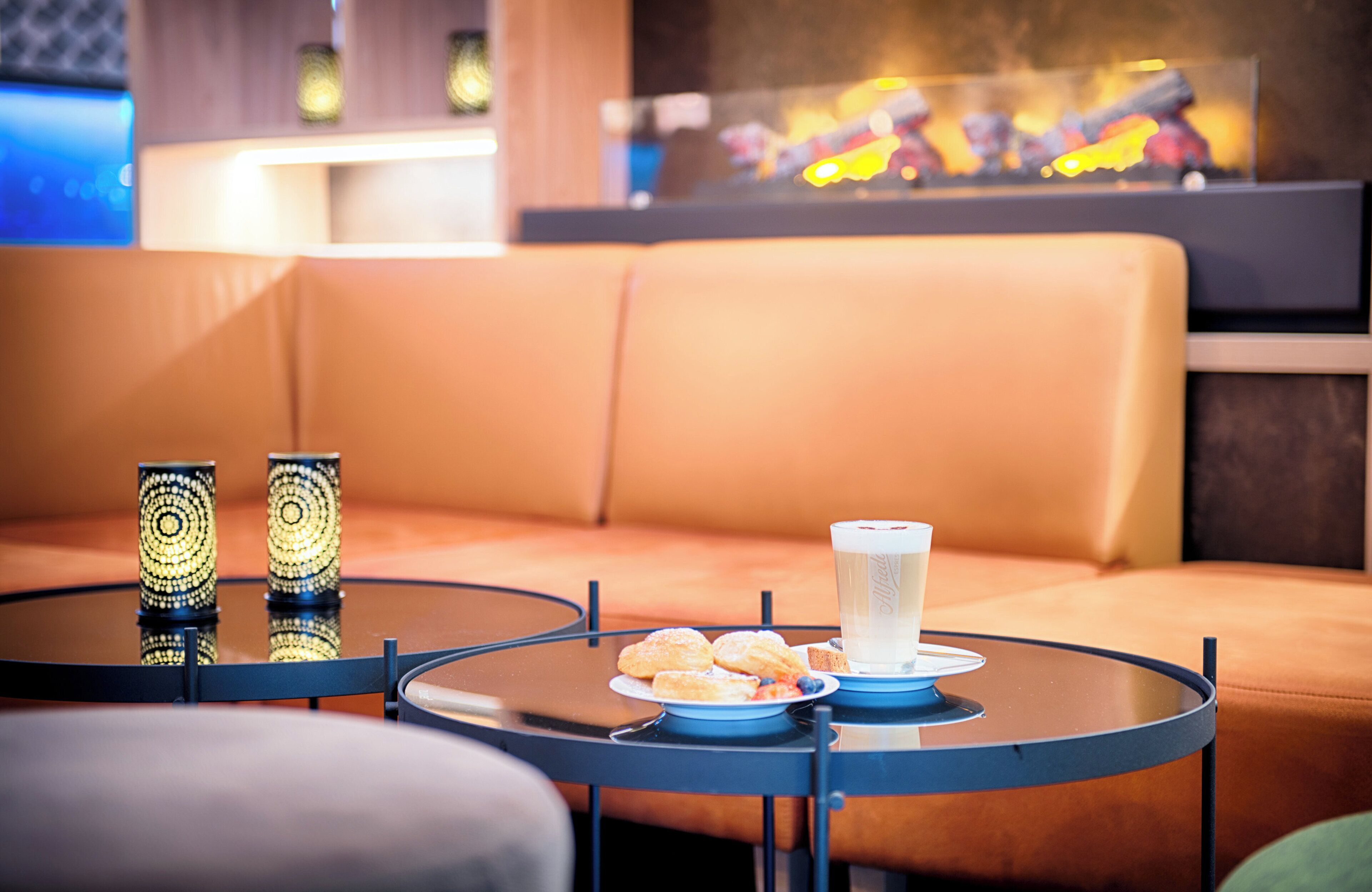 Un espace détente moderne avec une table ronde en verre, une assiette de pâtisseries, un verre d’eau et des bougies décoratives.