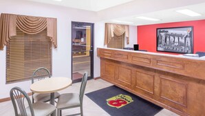Une chambre d’hôtel avec un comptoir d’accueil en bois, une table ronde entourée de chaises et une grande fenêtre avec des stores.