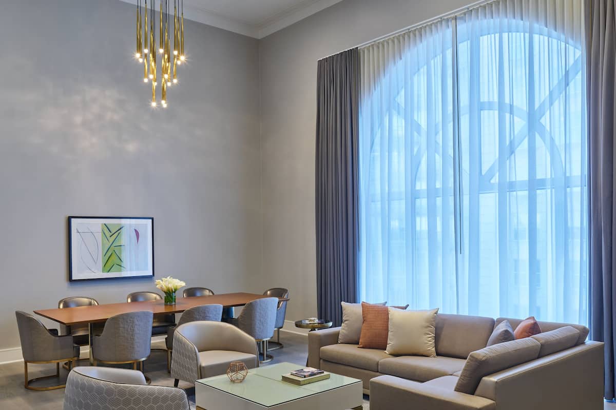 Ein modernes Wohnzimmer mit einer Couch, Hocker, Couchtisch und Sesseln. Es gibt einen Esstisch mit Stühlen und eine Deckenleuchte.