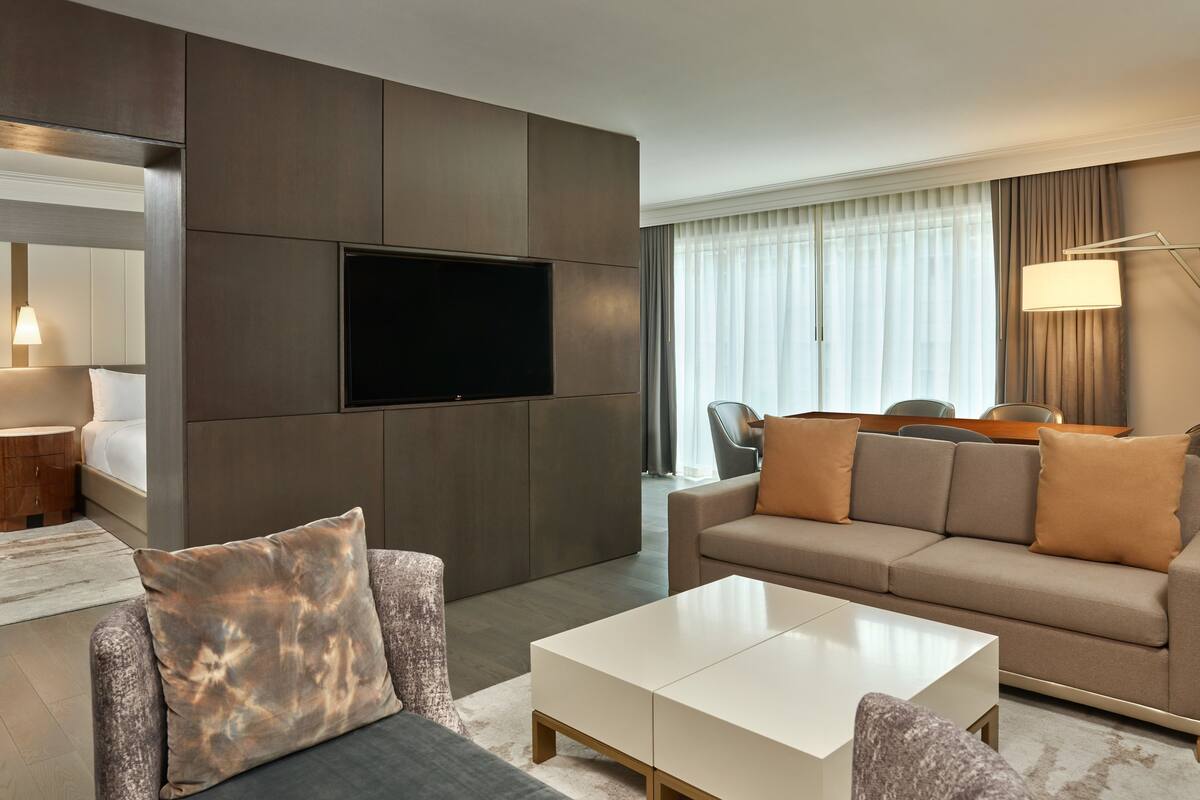 Ein modernes Hotelzimmer mit Flachbildfernseher an einer dunkelgrauen Wand, einem beigen Sofa mit passenden Kissen, einem weißen Couchtisch und einer Essecke mit Tisch und Stühlen.