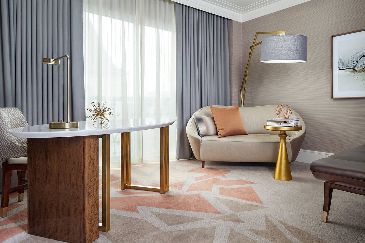 Ein modernes Hotelzimmer mit einem runden Esstisch, einer Couch, einem Beistelltisch mit Lampe und einem großen Fenster mit Vorhängen.