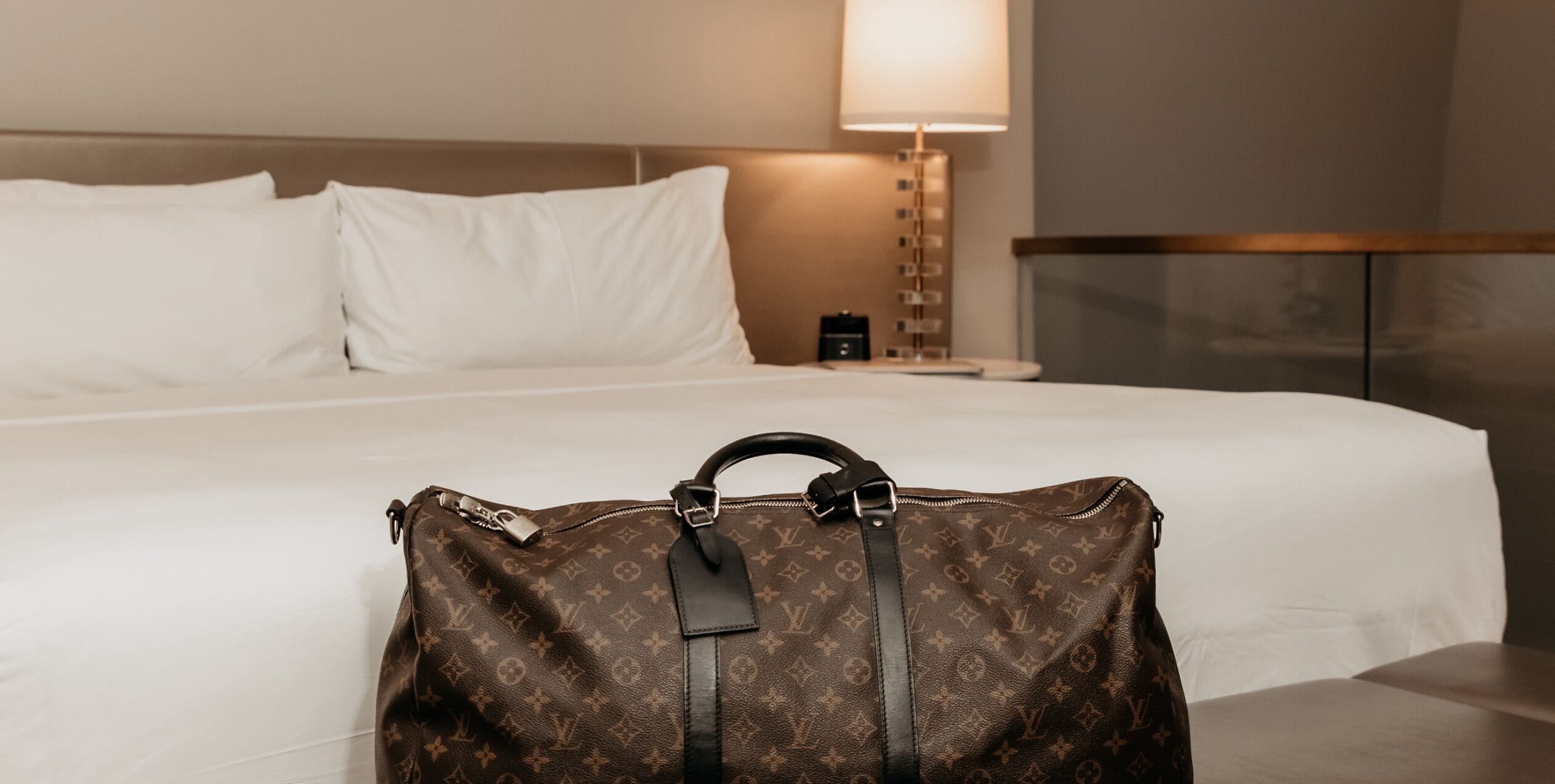 A bed with white linens, a brown Louis Vuitton bag on the bed, a bedside lamp, and a nightstand with a clock.