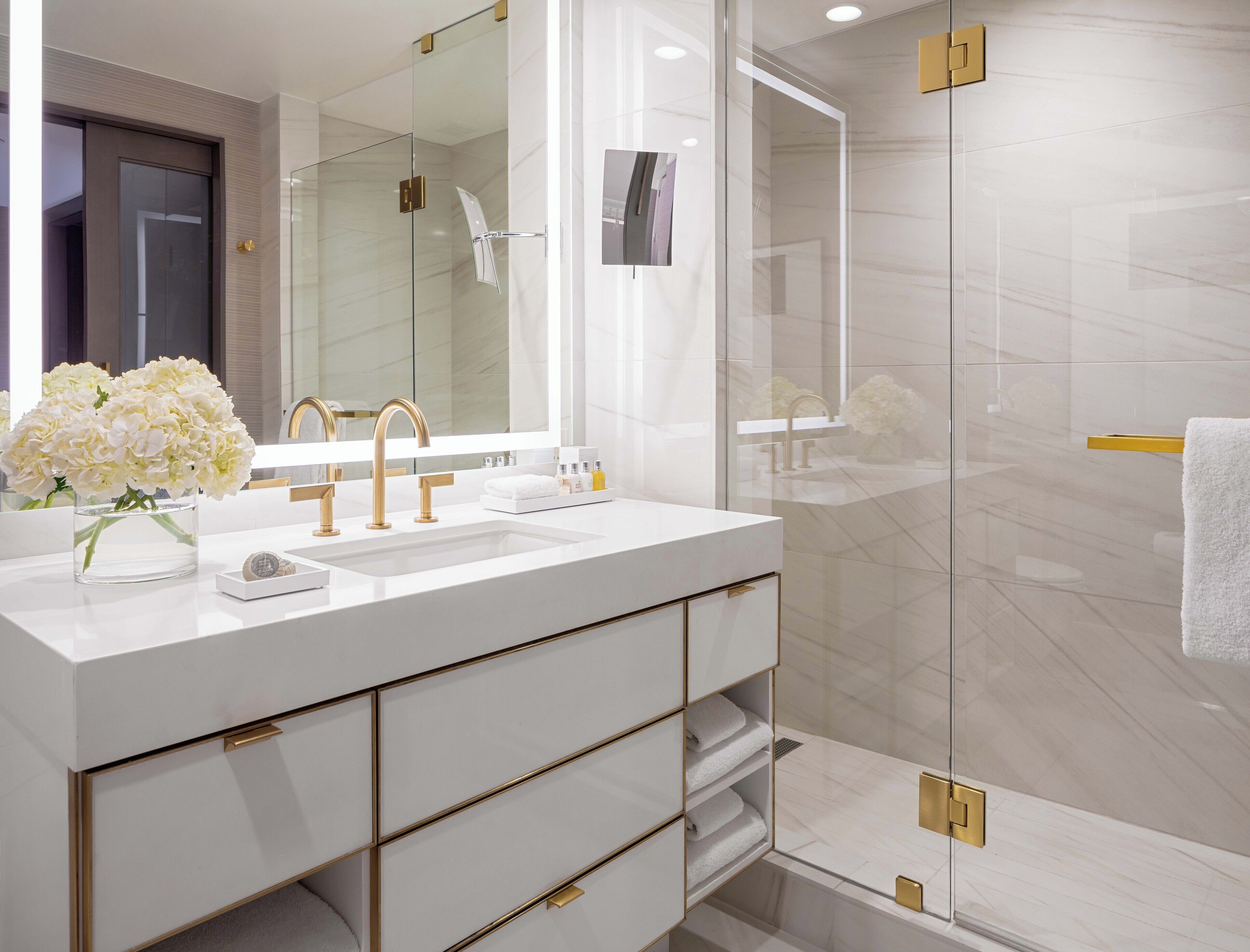 A modern bathroom with a large mirror, white sink, gold fixtures, and a glass shower enclosure.