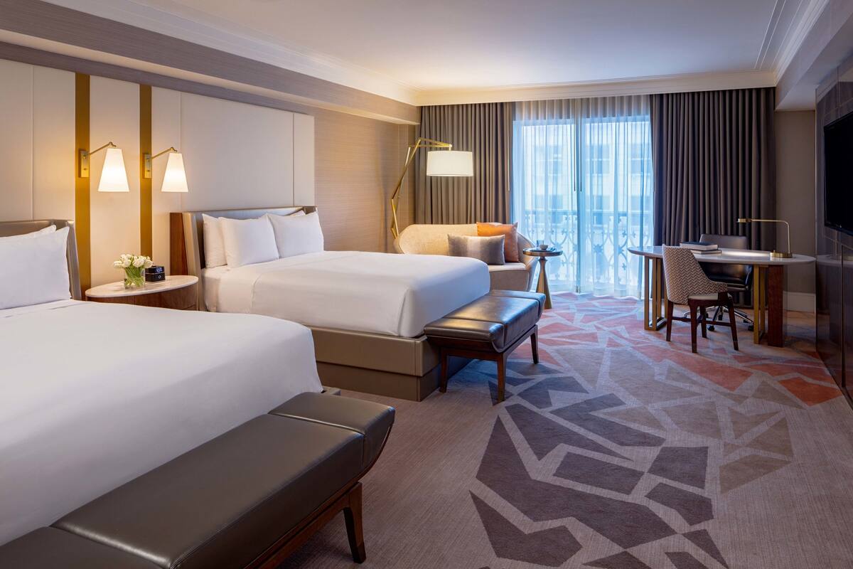 Ein Hotelzimmer mit zwei Betten, einem Schreibtisch, einem Sessel, einem Fernseher und einem großen Fenster mit Vorhängen.