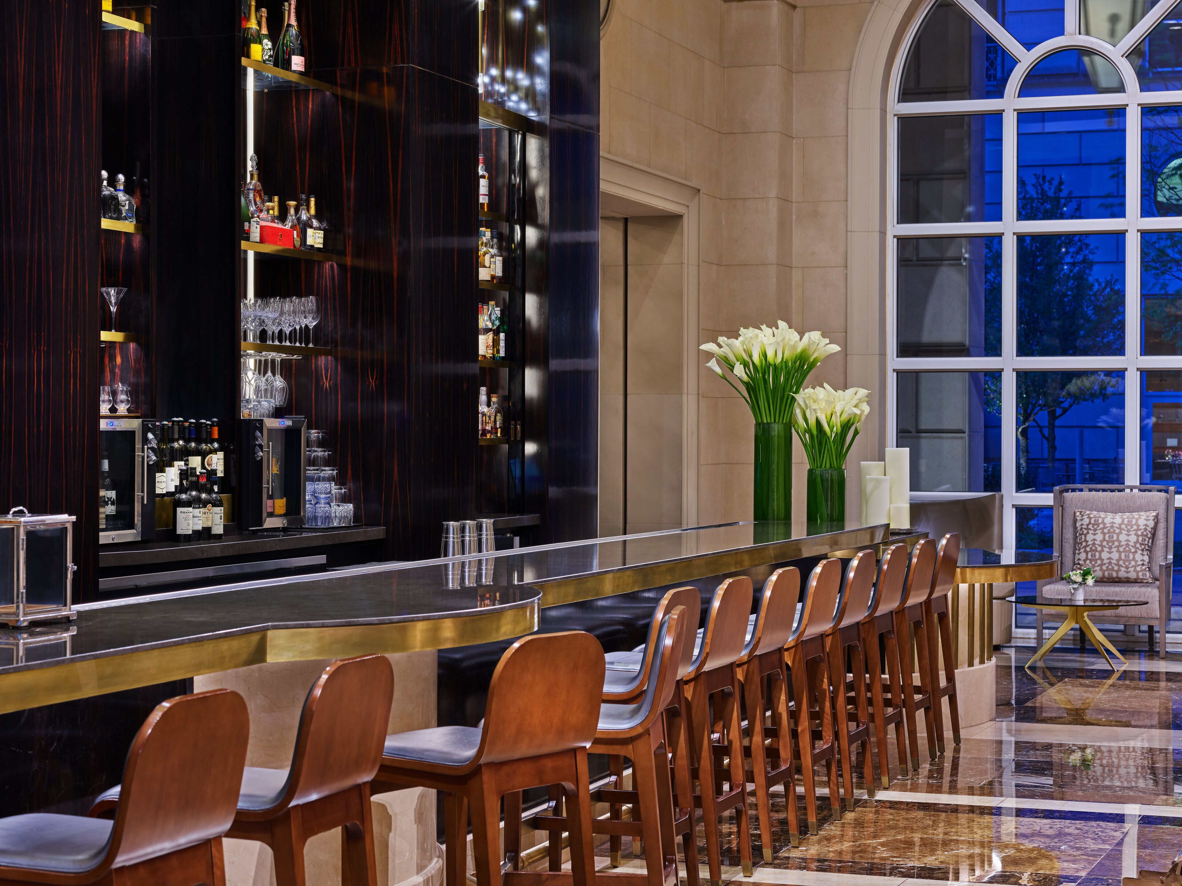 A bar area with a long counter, multiple shelves of bottles, and a row of wooden chairs.