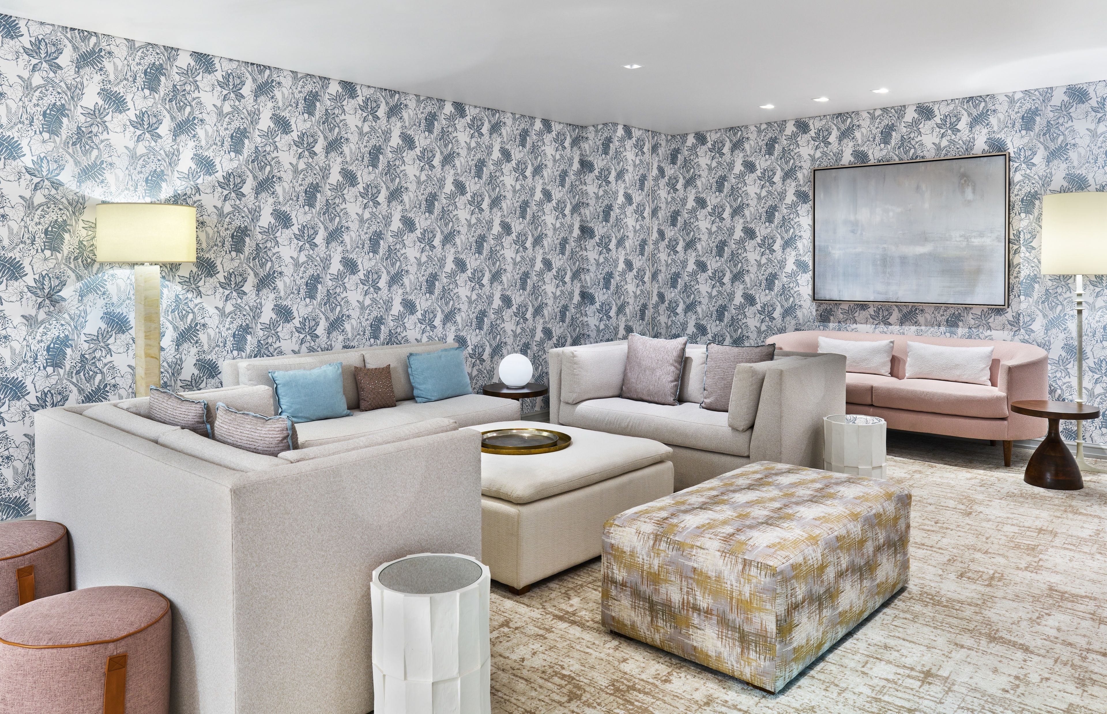 Ein Wohnzimmer mit Ecksofa, Hocker und Couchtisch. Zwei Lampen, eine Stehlampe und ein gerahmtes Kunstwerk sind an der Wand zu sehen.