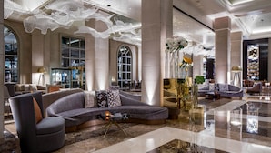 A spacious lobby with a curved grey sofa, a round armchair, and a coffee table. There are large columns, a chandelier, and a large window with a view of the city. - Hotel Crescent Court (Dallas)