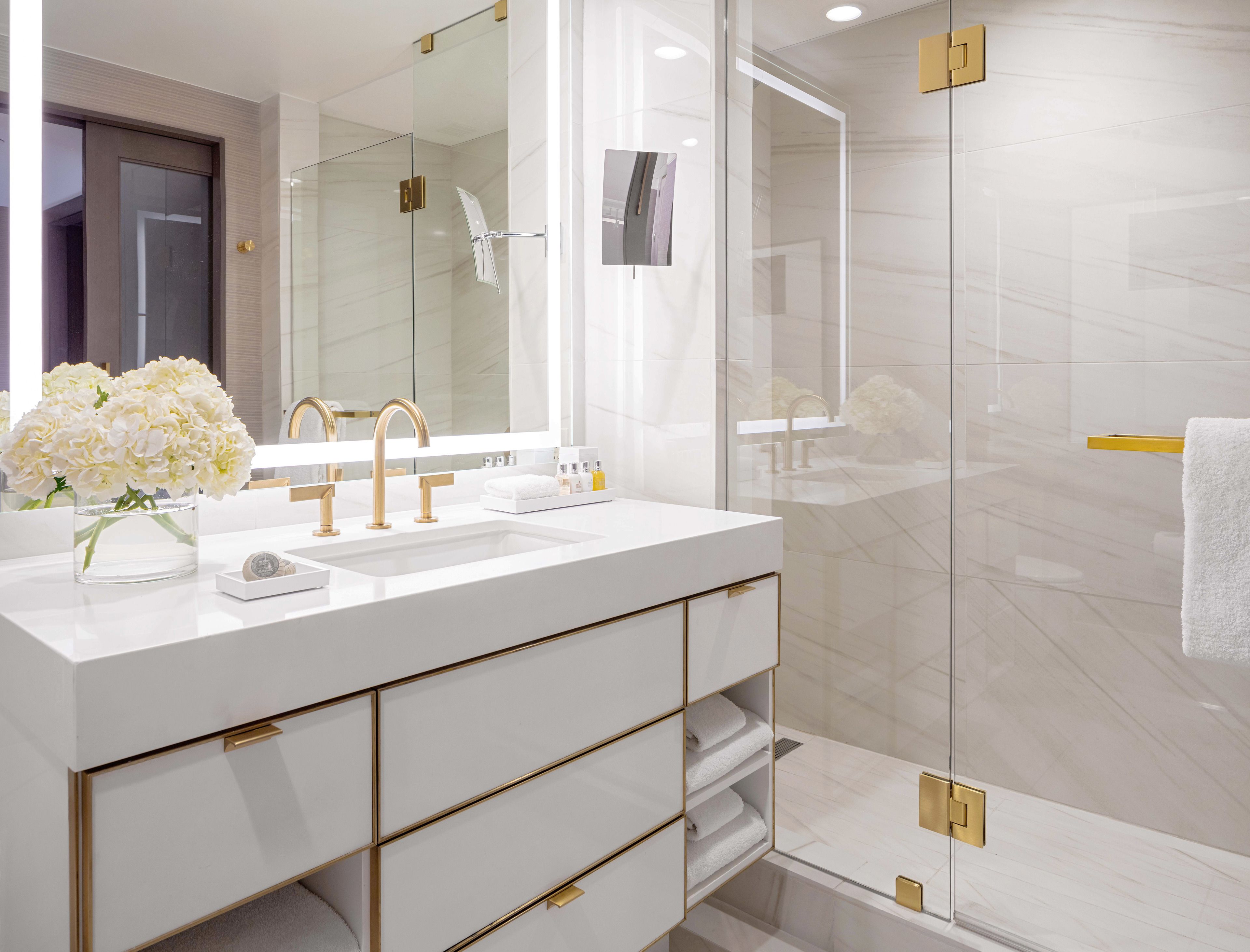 A modern bathroom with a white vanity, gold fixtures, and a glass shower enclosure.