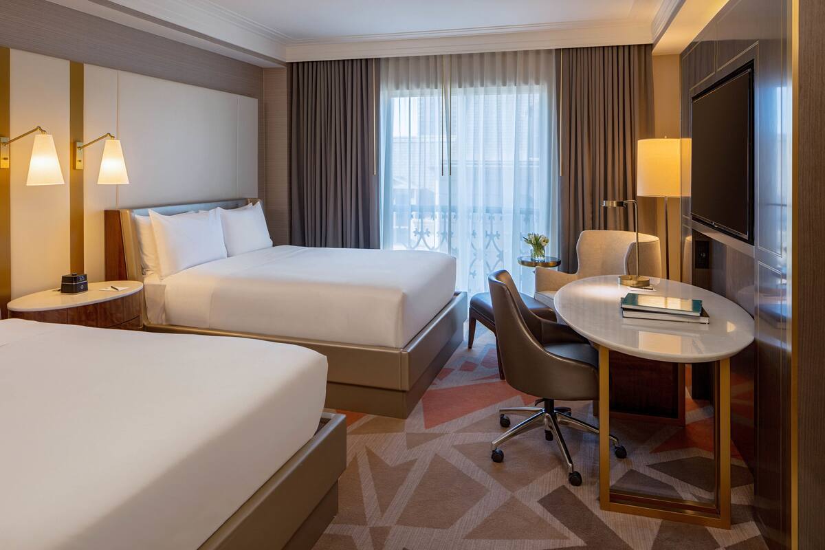 Ein Hotelzimmer mit zwei Betten, einem Schreibtisch mit Stuhl, einem Fernseher und einem Fenster mit Vorhängen.