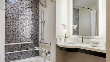 A modern bathroom with a glass mosaic shower, a white bathtub, a vanity with a mirror, and a potted orchid.