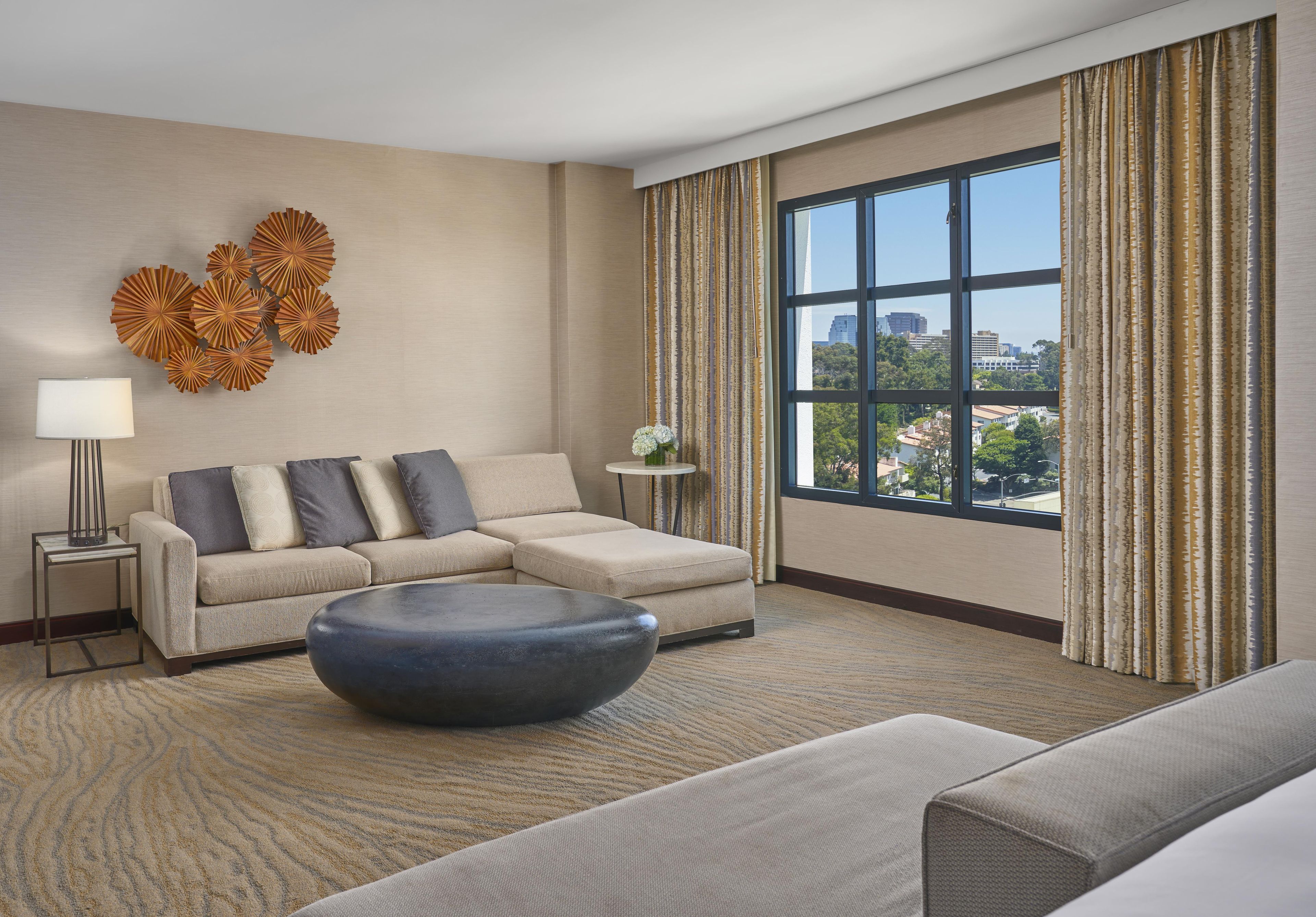 A hotel room with a sectional sofa, a round ottoman, a side table with a lamp, a decorative wall piece, and a large window with curtains.