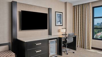 A hotel room with a flat-screen TV, a desk with a chair, a small table with a lamp, a framed picture on the wall, and a large window with curtains.