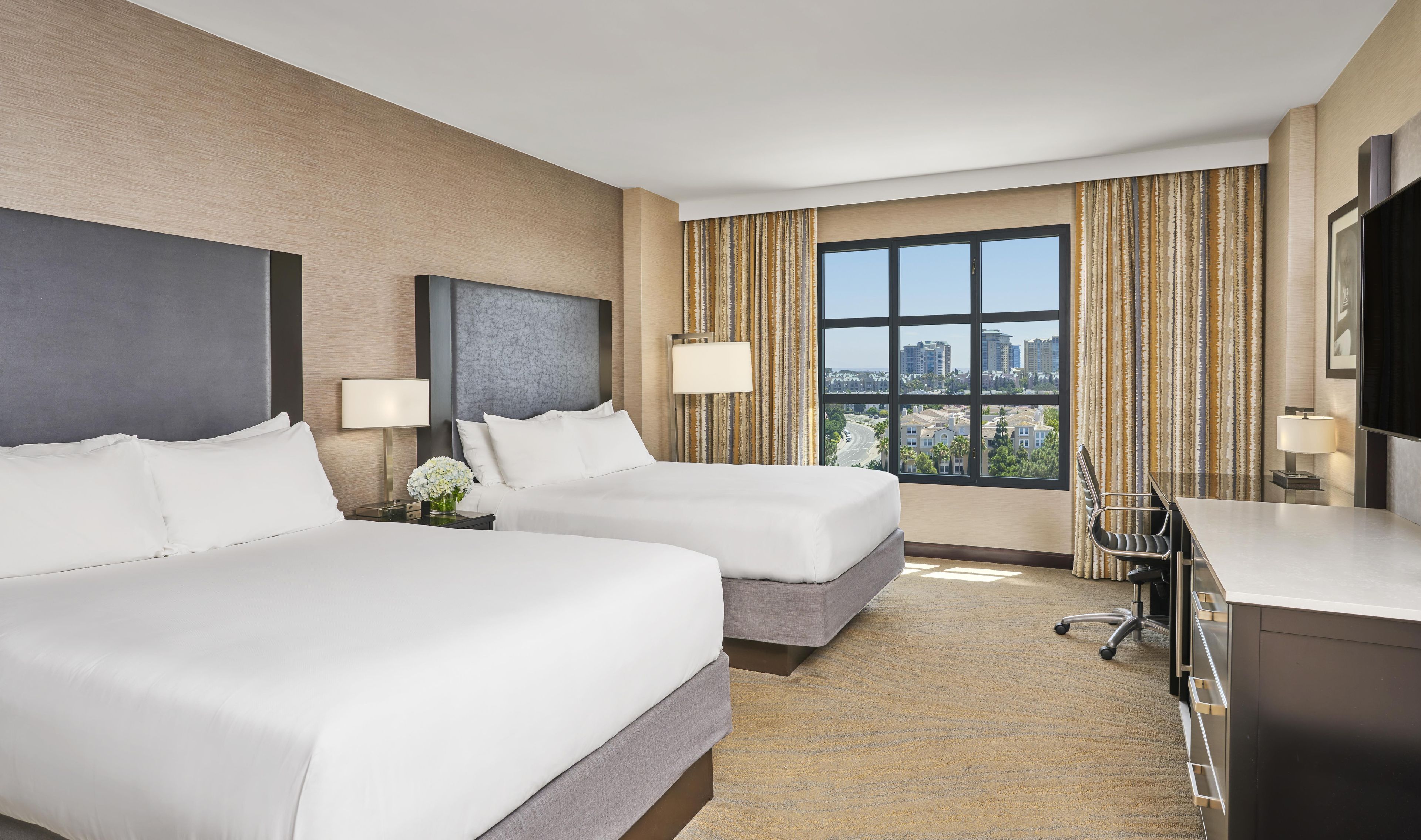 A hotel room with two beds, a desk, a chair, and a large window with a city view.