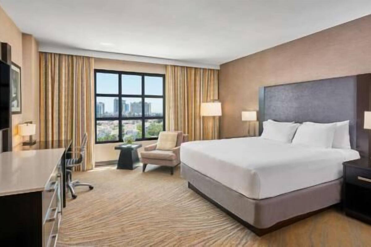 A hotel room with a large bed, a desk, a chair, a TV, and a window with curtains.
