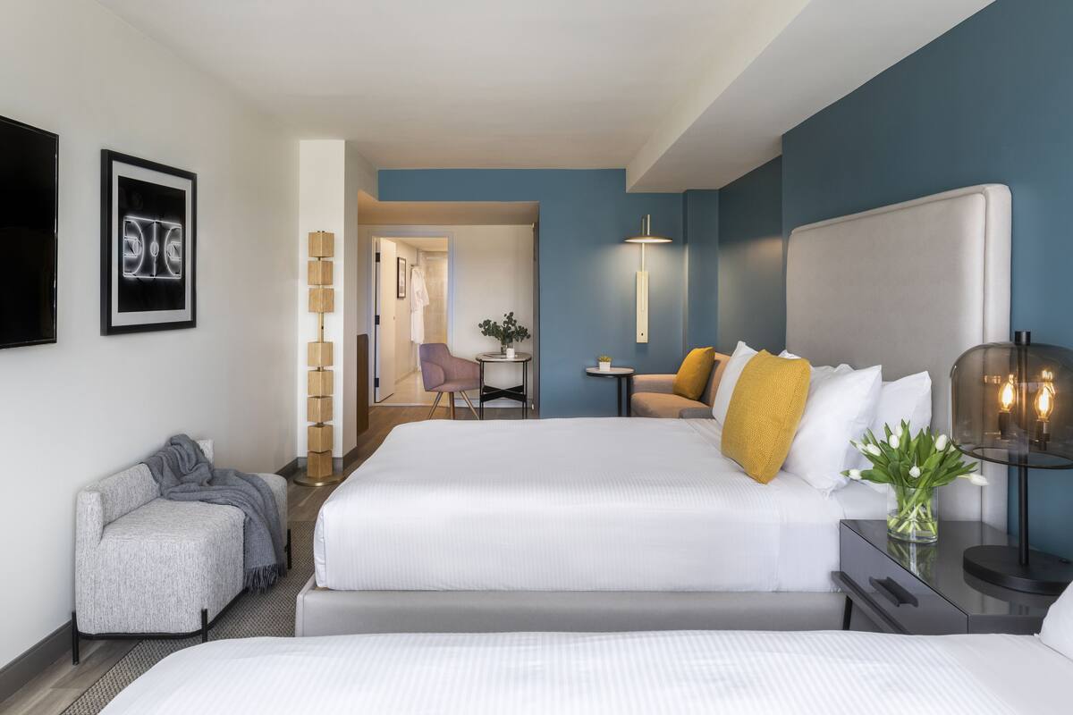 A hotel room with two beds, a small seating area, a desk, and a view of a hallway.
