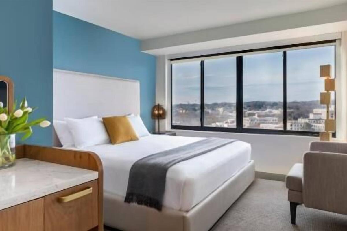 A hotel room with a bed, a desk, a chair, a vase with flowers, and a large window with a city view.