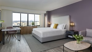 A modern hotel room with a large bed, a desk, a chair, a small table, and a view of the city through large windows. - Hotel Madera (Washington)