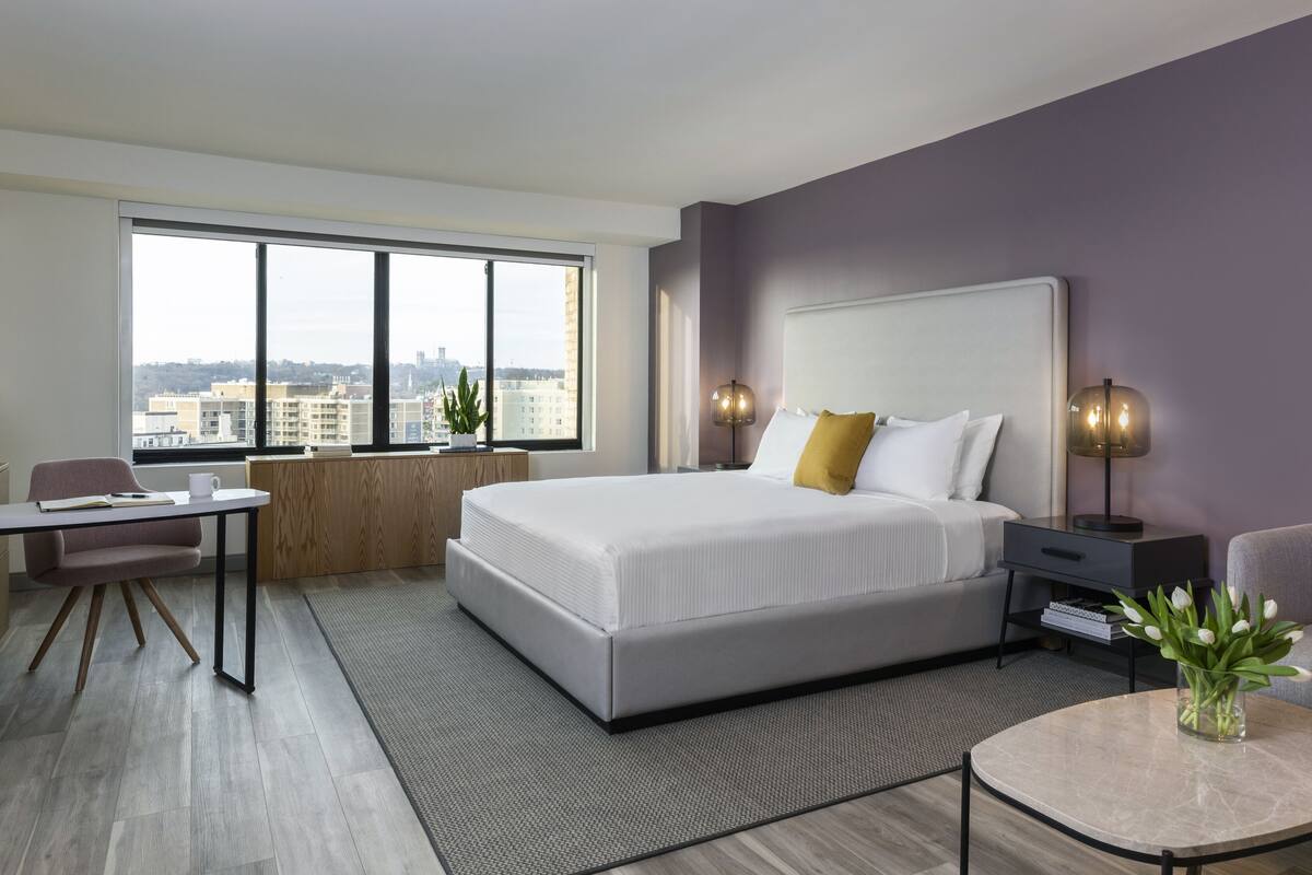 A modern hotel room with a large bed, a desk, a chair, a small table, and a view of the city through large windows.