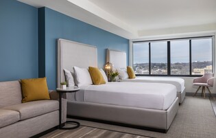 A modern hotel room with two beds, a sofa, a small table, and a chair. The room has a large window with a city view.
