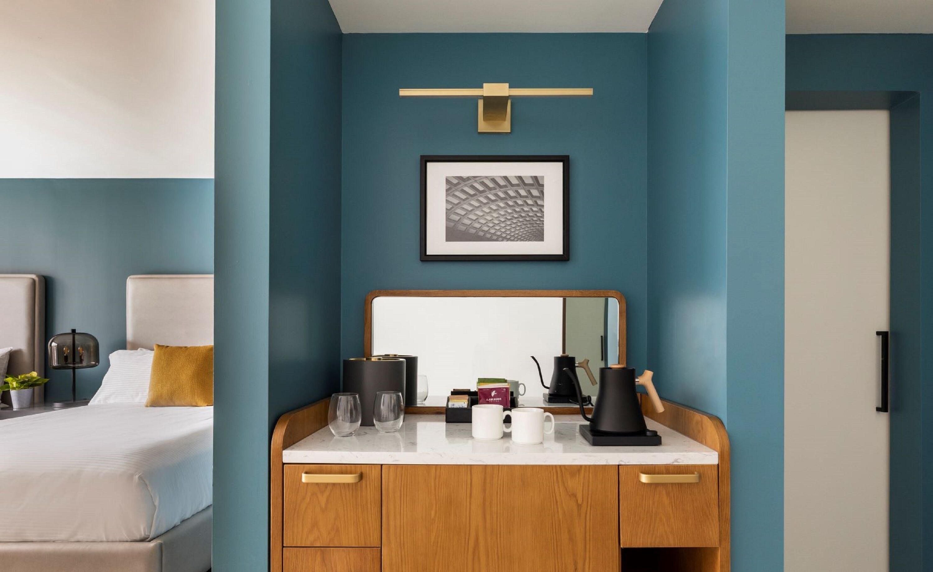 A modern hotel room with a teal accent wall, a large mirror, a wooden cabinet with a coffee machine, and a framed picture above the mirror.