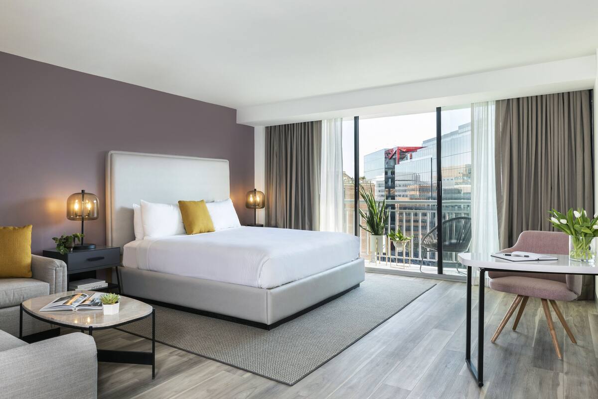 A modern hotel room with a large bed, a sofa, a small table, and a balcony with a view of the city.