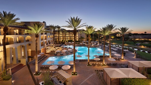 A hotel with a large outdoor pool surrounded by palm trees and umbrellas, multiple floors with balconies, and a well-lit evening setting. - Fairmont Scottsdale Princess (Scottsdale)