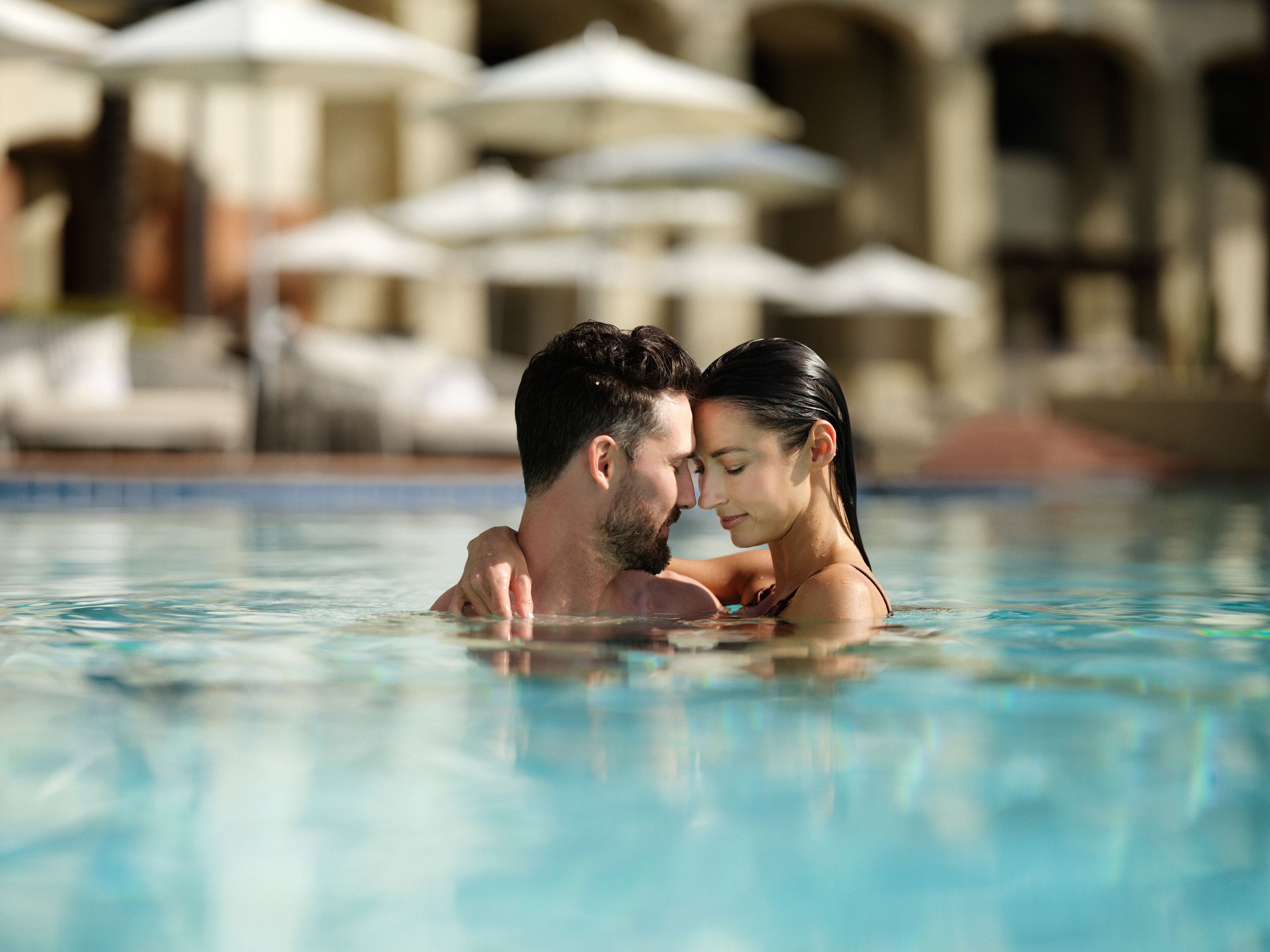 A couple in a swimming pool with a building and umbrellas in the background.