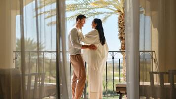 Una pareja abrazándose en un balcón con vista a palmeras y un cielo despejado.