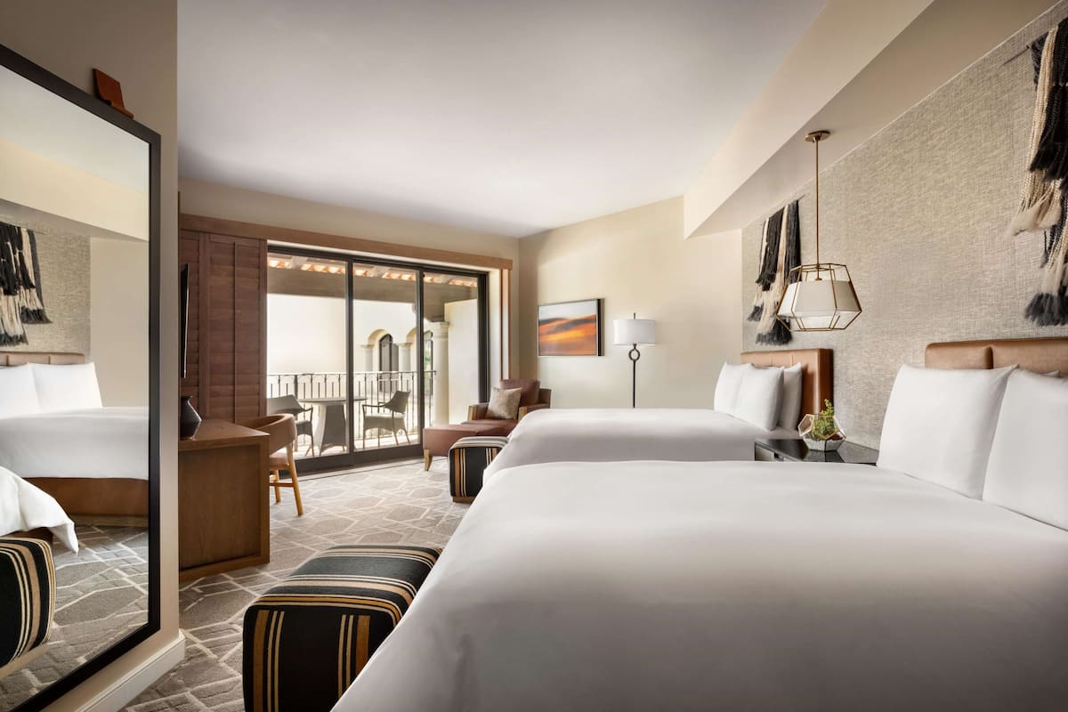 Ein Hotelzimmer mit zwei Betten, einem Schreibtisch, einem Stuhl, einer Couch, einer Lampe und einem großen Fenster mit Blick auf ein Gebäude.