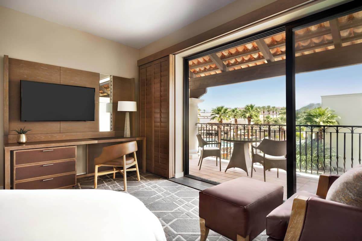 Ein Hotelzimmer mit Bett, Schreibtisch, Fernseher, einem Balkon mit Stühlen und Tisch sowie Blick auf Palmen.