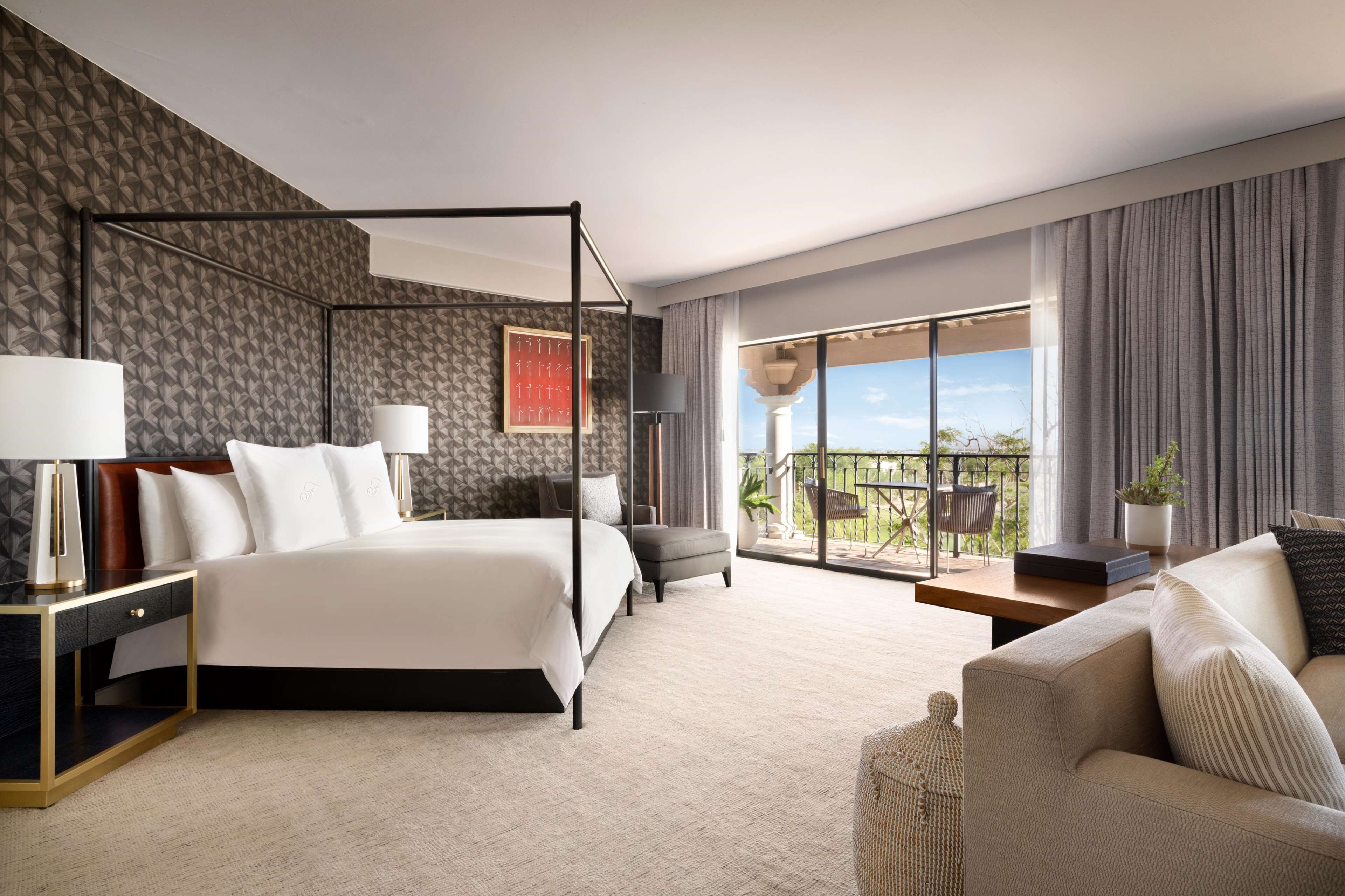 A modern hotel room with a four-poster bed, a balcony with a view, and a seating area with a sofa and armchair.