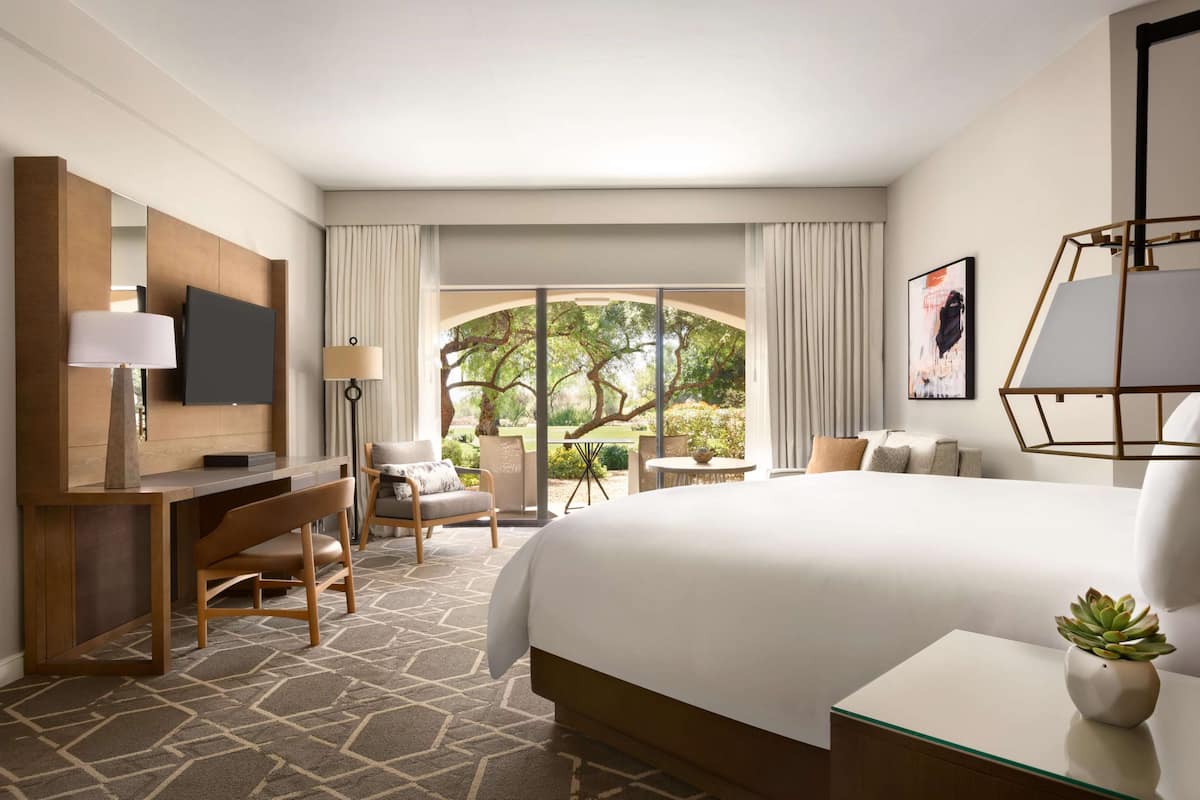 Ein Hotelzimmer mit einem großen Bett, einem Schreibtisch mit Stuhl, einem Fernseher und Blick auf einen Garten durch ein großes Fenster.