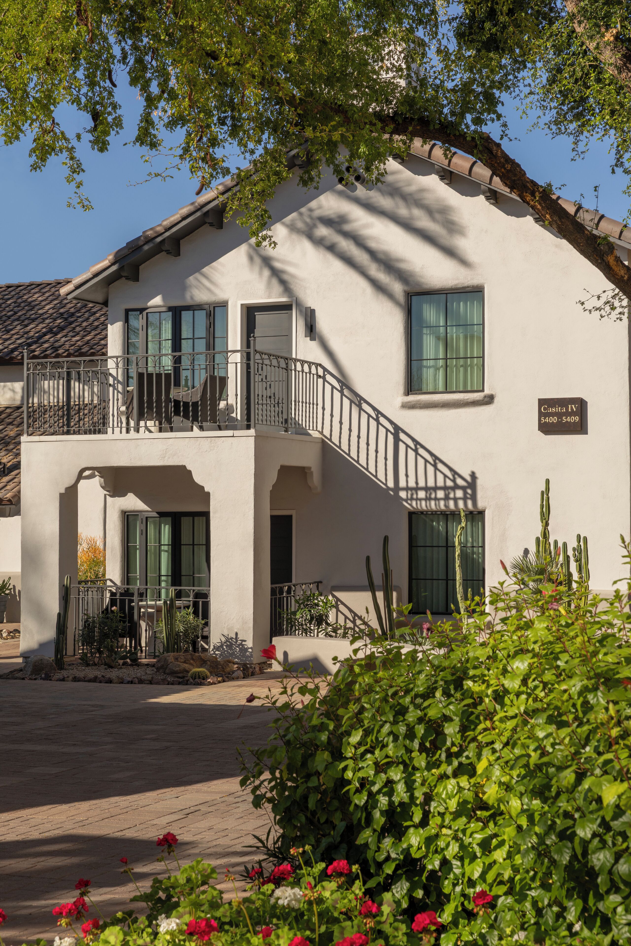 A two-story building with a balcony, a sign indicating 'Casita IV 5400-5409', and a courtyard with plants and flowers.